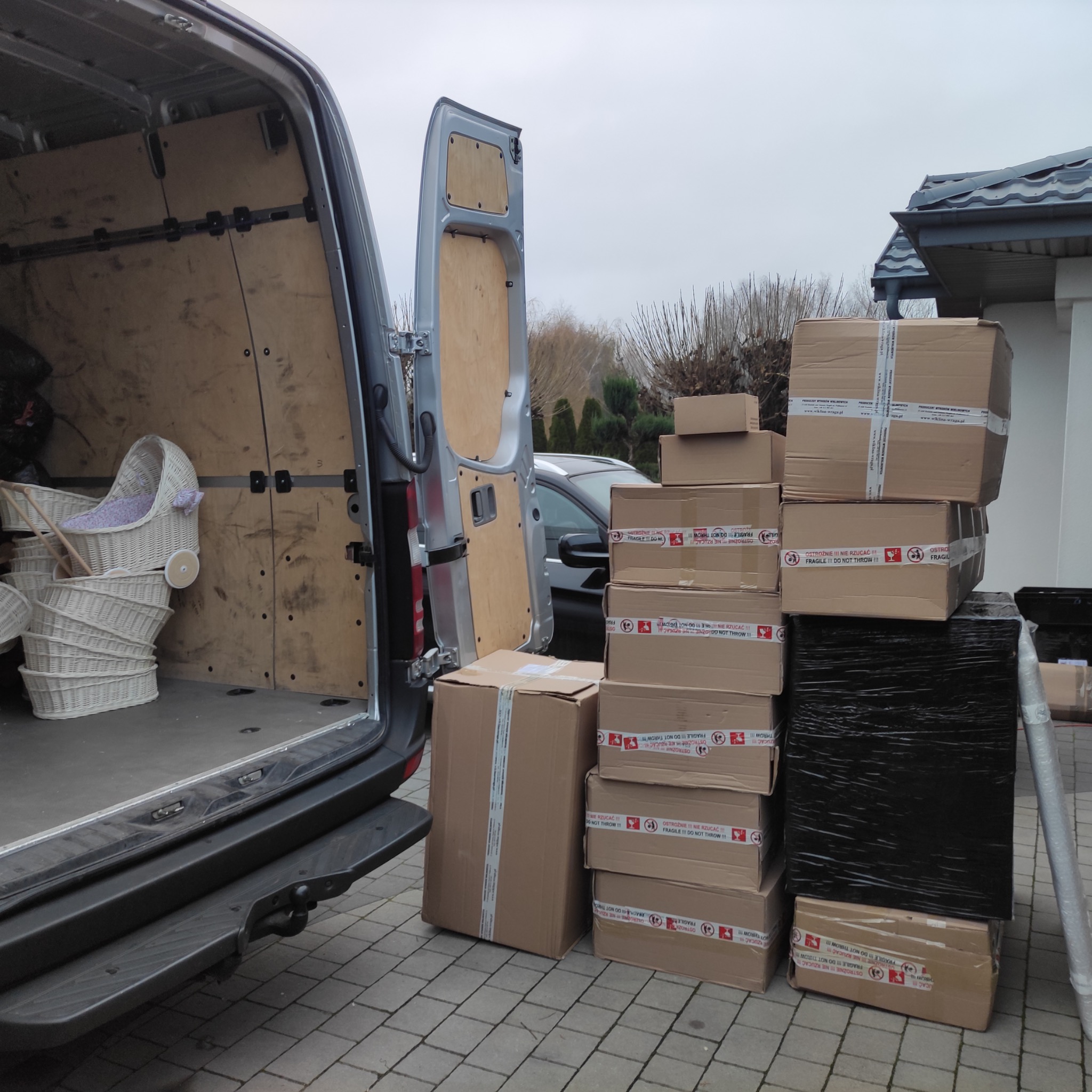 Częściowo załadowany bus dostawczy z otwartymi drzwiami, obok sterta zapakowanych kartonów z napisem 'Fragile' i wiklinowe kosze wewnątrz, na zewnątrz kostka brukowa i dom z szarą dachówką.