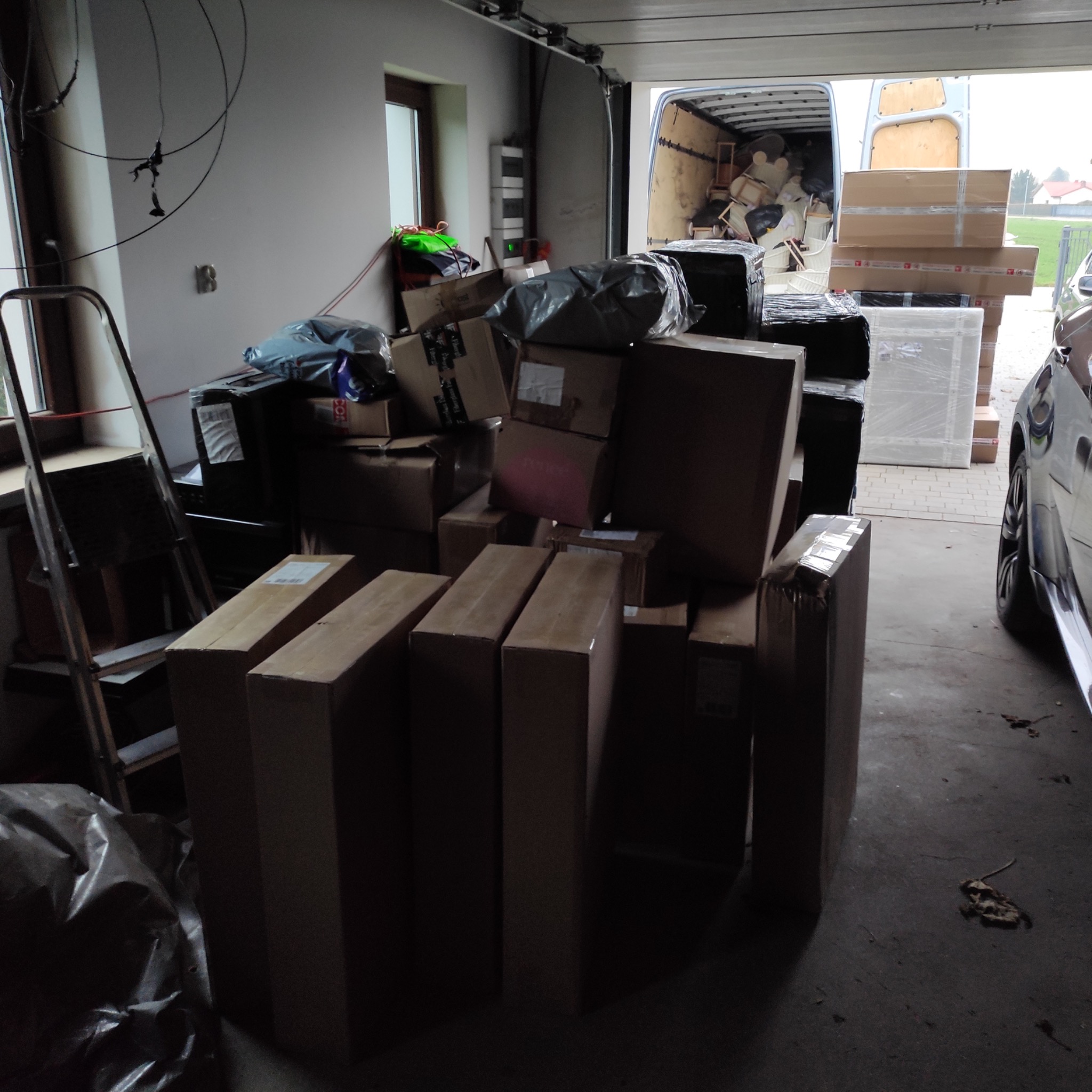 Stos kartonowych pudeł i zapakowanych przedmiotów w garażu, częściowo załadowanych do furgonetki, widoczna drabina i fragment samochodu.