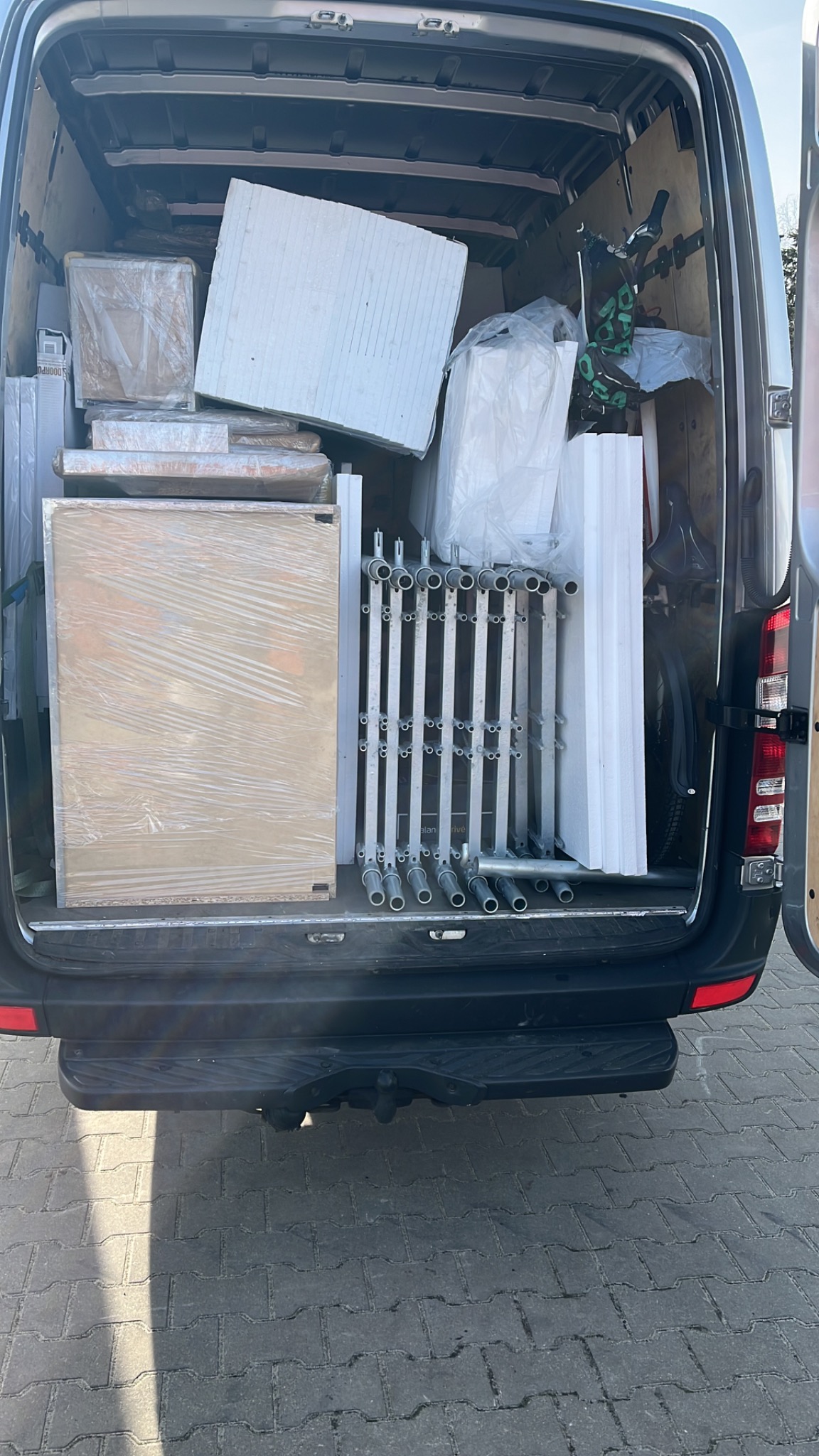 Wnętrze furgonetki załadowane elementami konstrukcyjnymi, płytami i opakowanymi przedmiotami, gotowe do transportu.