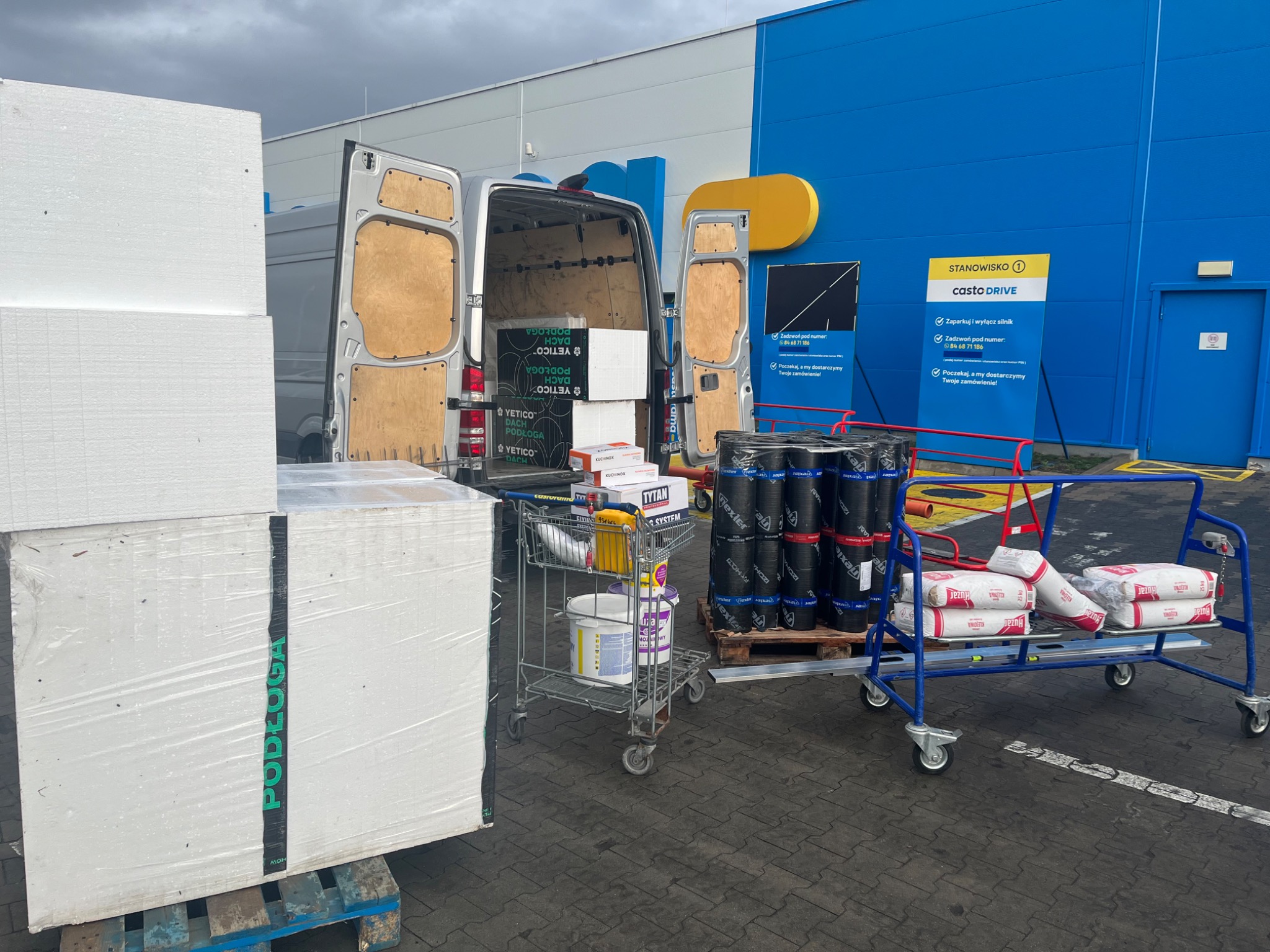 Załadunek materiałów budowlanych do busa przy stanowisku Casto Drive: styropian na palecie z napisem PODŁOGA, opakowania dachowe YETICO, farby, kleje i worki z zaprawą na wózkach transportowych.