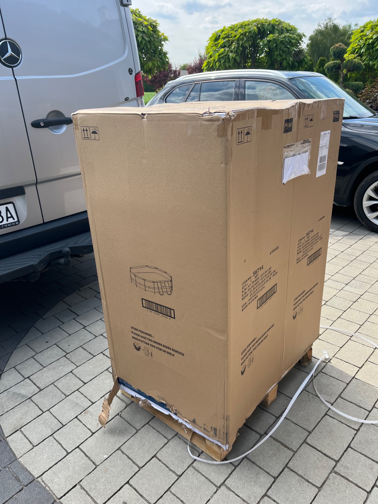 Duże kartonowe pudło z nadrukowanym schematem basenu ogrodowego na palecie, zabezpieczone taśmami transportowymi, obok zaparkowany srebrny van Mercedes i czarny samochód osobowy na brukowanym...