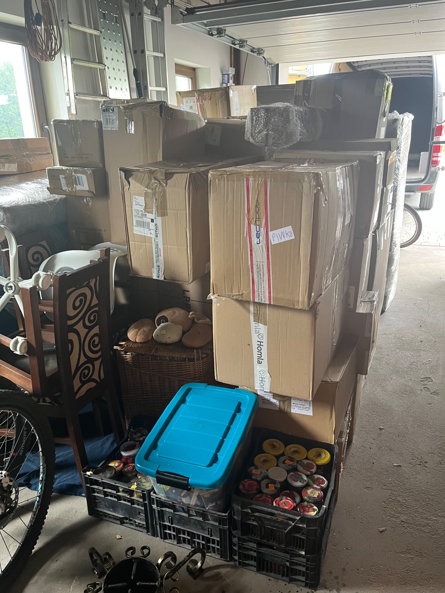 Stos kartonowych pudeł z zapakowanymi przedmiotami, ustawionych w garażu, przygotowanych do załadunku do furgonetki, widoczna skrzynka z przetworami i niebieski pojemnik.