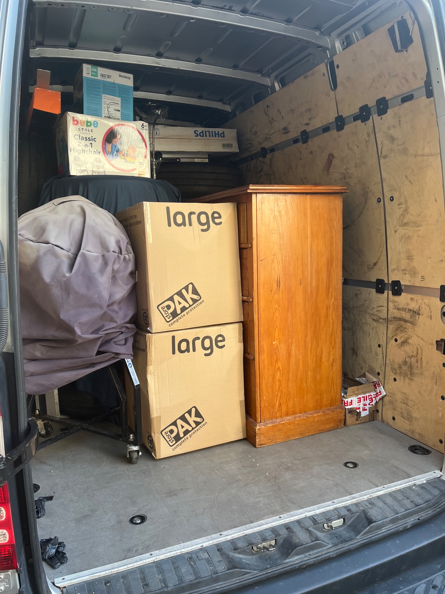 Wnętrze furgonetki załadowane kartonami z napisem 'large', meblami, pudłami z zabawkami i akcesoriami dziecięcymi, przygotowanymi do transportu.