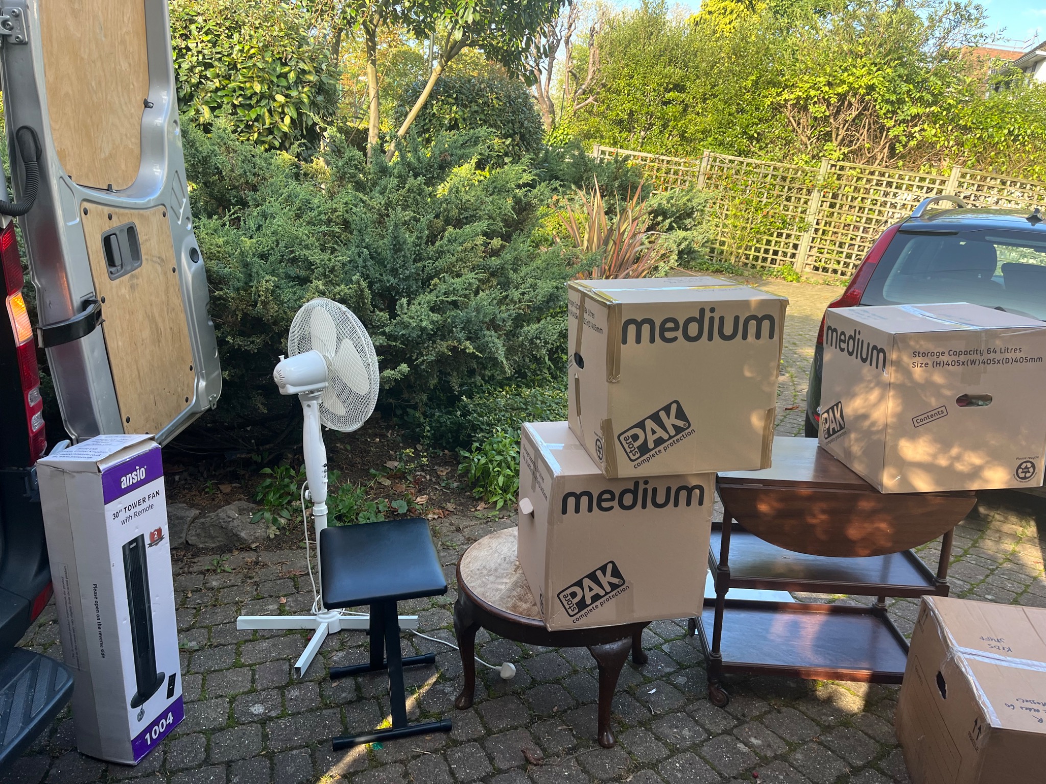 Przygotowania do transportu: otwarte drzwi furgonetki, kartony z napisem 'medium' i logo 'PAK complete protection', wentylator stojący, mały stołek, stolik na kółkach i pudełko wentylatora...