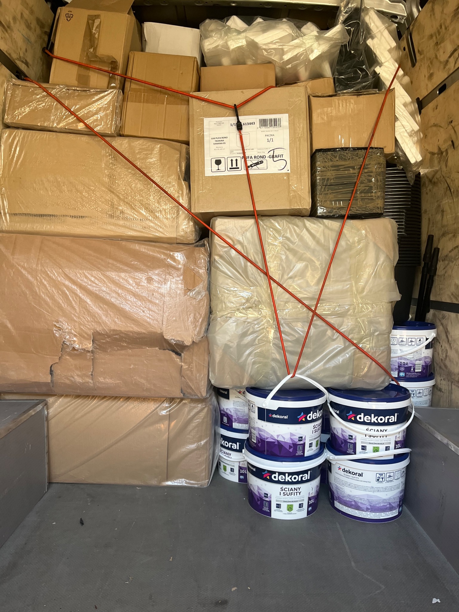 Wnętrze furgonetki załadowane kartonami, folią i wiaderkami z farbą Dekoral, zabezpieczone czerwonymi gumami transportowymi.