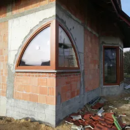 Narożnik budynku w trakcie budowy z zamontowanym półokrągłym oknem z brązową ramą i prostokątnym oknem w tle, widoczne cegły, beton i elementy rusztowania.