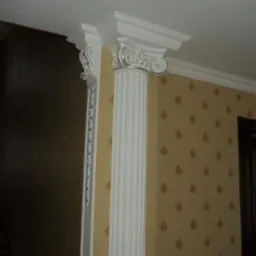 Biała kolumna dekoracyjna z kapitelem jońskim, zamontowana przy ścianie z wzorzystą tapetą w odcieniach beżu i brązu, widoczny fragment schodów w tle.