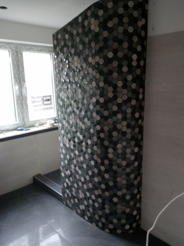 Ścianka prysznicowa wykończona mozaiką heksagonalną w odcieniach szarości i czerni, widoczna z perspektywy podłogi, z oknem w tle.