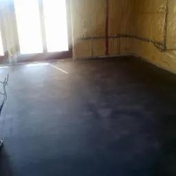 Wnętrze w trakcie wykańczania z gładką, ciemną wylewką na podłodze, widoczne przewody elektryczne przy ścianach i jasne światło wpadające przez okno.