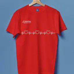 Czerwona koszulka z białym nadrukiem: logo 'Stanley Centrum Medyczne' i linia EKG z trzema sercami, wisząca na drewnianym wieszaku na tle błękitnej ściany.