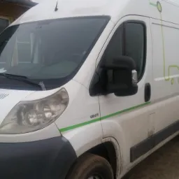 Biały samochód dostawczy Peugeot z zielonymi akcentami i grafiką stylizowanego auta, zaparkowany na błotnistym terenie przed budynkiem z oknami, w pochmurny dzień.