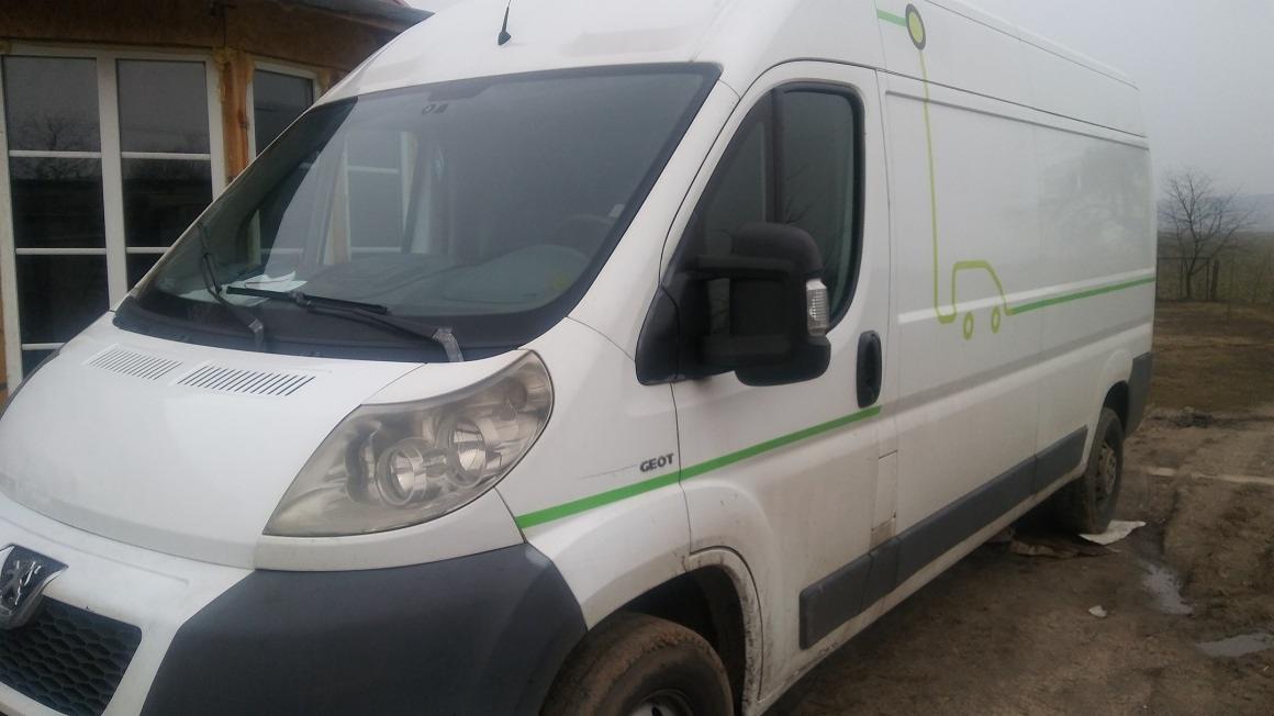 Biały samochód dostawczy Peugeot z zielonymi akcentami i grafiką stylizowanego auta, zaparkowany na błotnistym terenie przed budynkiem z oknami, w pochmurny dzień.