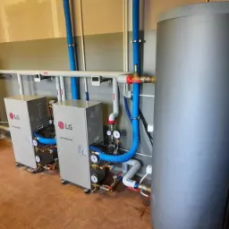 Dwa identyczne moduły grzewcze LG Thermav V z podłączonymi rurami, zbiornik buforowy i elementy instalacji hydraulicznej w pomieszczeniu z terakotową podłogą i szarą lamperią.