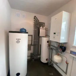 Kompaktowa kotłownia w Ryczówku z białym zasobnikiem Kospel, kotłem gazowym Panasonic i szarymi rurami instalacyjnymi.