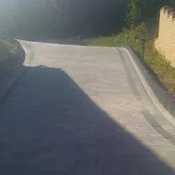 Szeroki, wybrukowany podjazd z szarej kostki na posesji, z żółtą ceglaną ścianą po prawej stronie i skarpą porośniętą zielenią po lewej, oświetlony silnym słońcem.