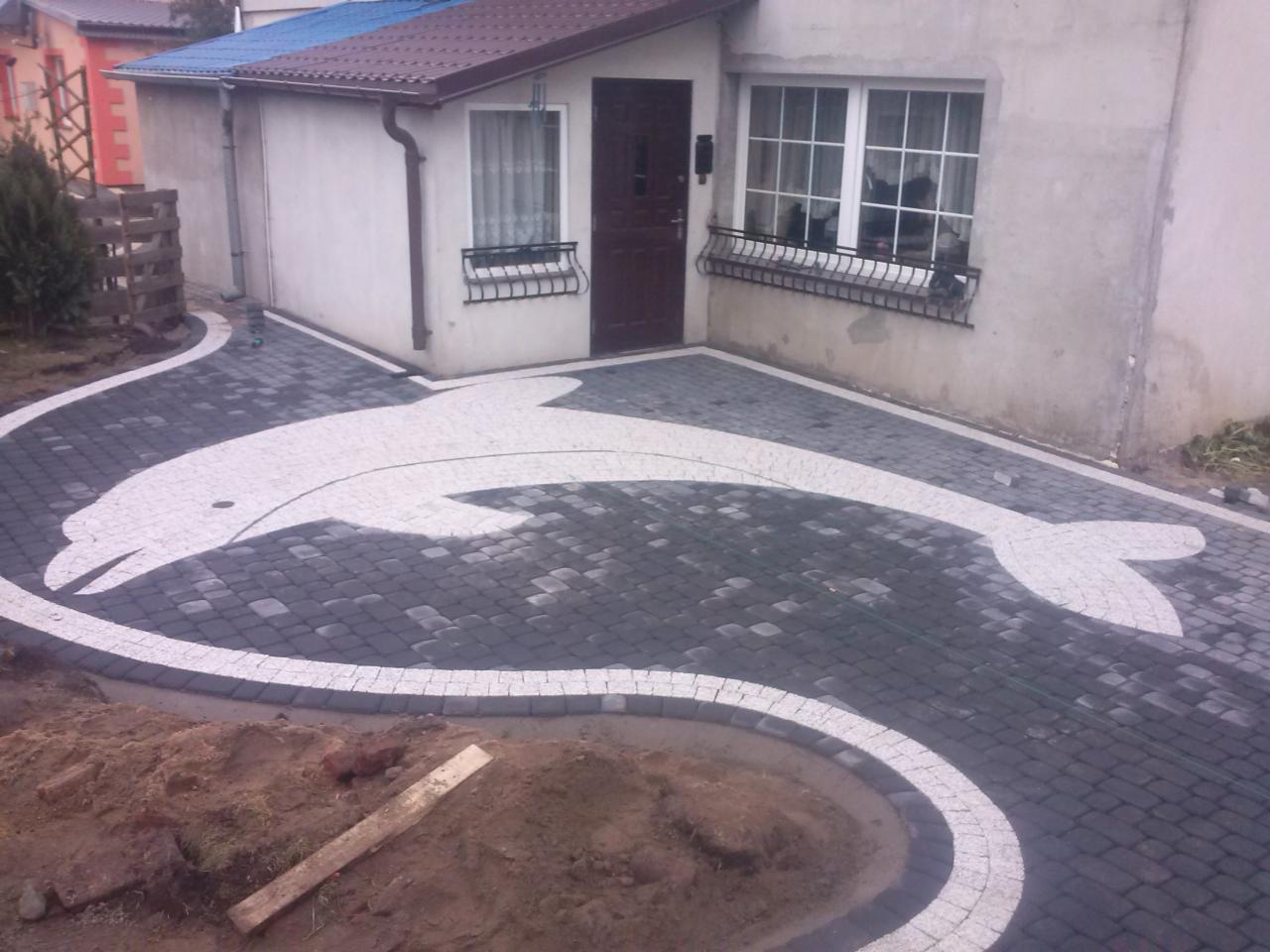 Podjazd z kostki brukowej ułożonej w kształt delfina, obrzeża z jasnej kostki, w tle dom jednorodzinny z brązowymi oknami.