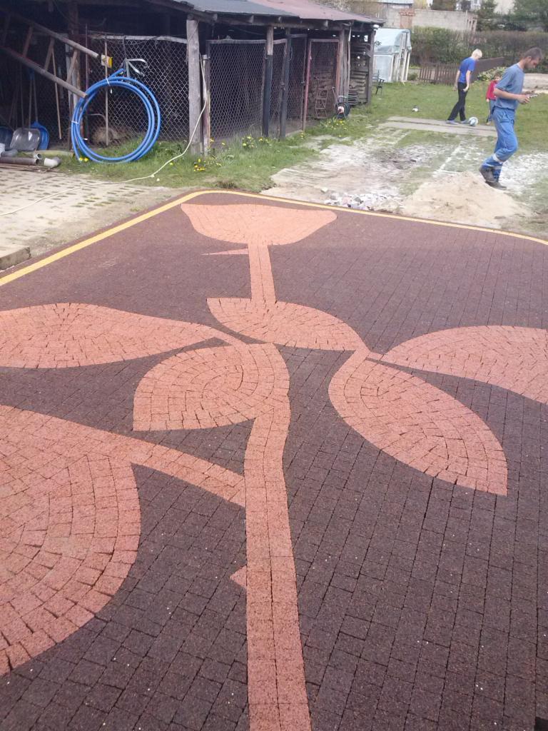 Ułożony wzór kwiatowy z kostki brukowej w kolorach brązu i beżu, obrzeże z żółtą linią, w tle budynek gospodarczy i osoby na trawniku.