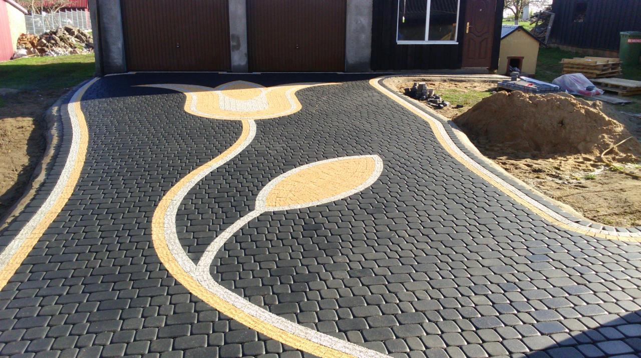 Czarno-szara kostka brukowa ułożona w kształt ścieżki z motywem kwiatowym w kolorze żółtym i białym, prowadząca do garażu.