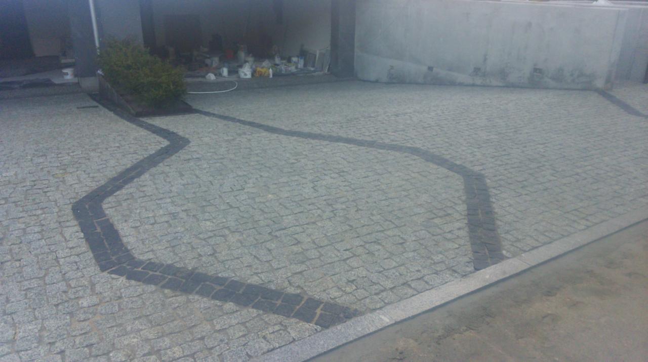 Ułożona kostka granitowa na podjeździe z ciemnoszarą obwódką tworzącą wzór zygzaka, mały krzew przy ścianie budynku, widoczne ślady po budowie.