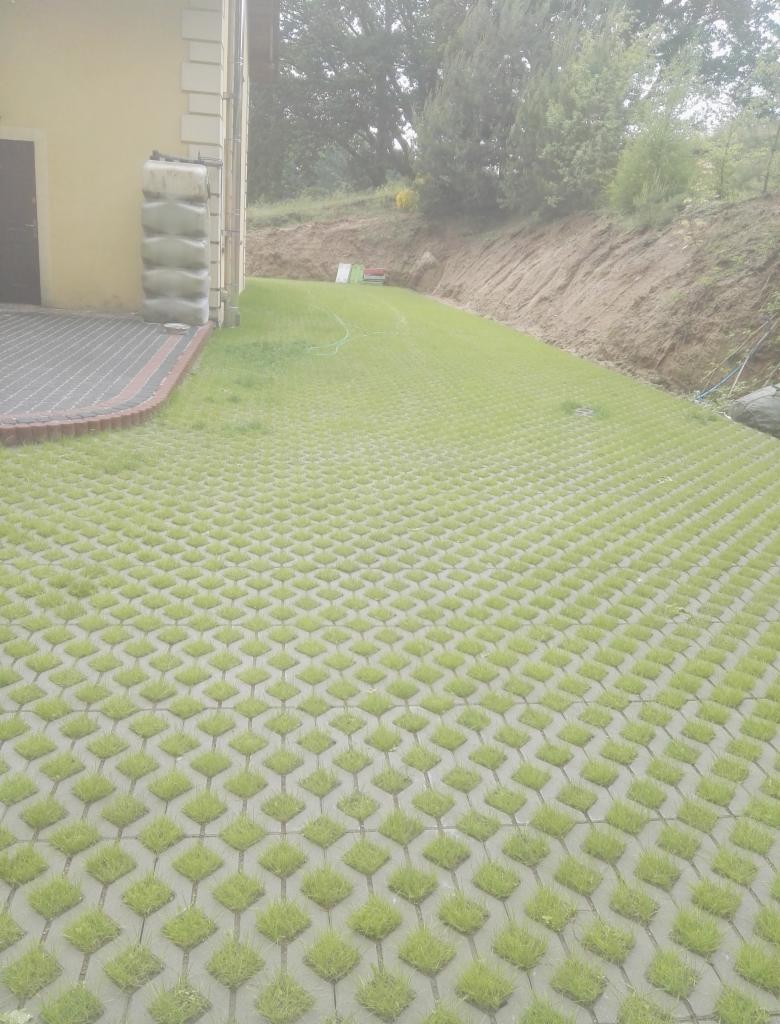 Eko-parking z ażurowych płyt betonowych wypełnionych trawą, przylegający do budynku z widocznym zbiornikiem na deszczówkę i fragmentem tradycyjnej kostki brukowej. W tle skarpa porośnięta zielenią.