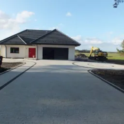 Nowoczesny dom z podjazdem z jasnoszarej kostki brukowej z ciemnymi obrzeżami; pracownik wykonuje prace ziemne przy krawędzi, w tle koparka na trawniku i srebrny samochód.