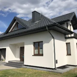 Nowoczesny dom jednorodzinny z białą elewacją, ciemną dachówką i grafitowymi elementami wykończeniowymi, widok z poziomu trawnika w słoneczny dzień.