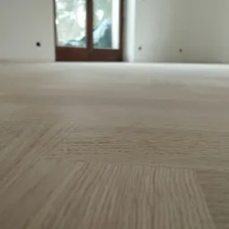 Jasna, nowo ułożona podłoga z paneli laminowanych z widocznym wzorem drewna, w tle drzwi balkonowe i ściany w stanie deweloperskim.