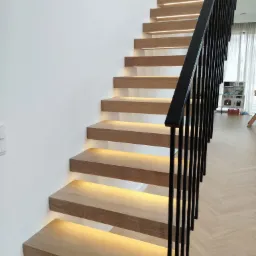 Nowoczesne schody z drewna z podświetleniem LED pod stopniami i czarną metalową balustradą, widok z dołu.