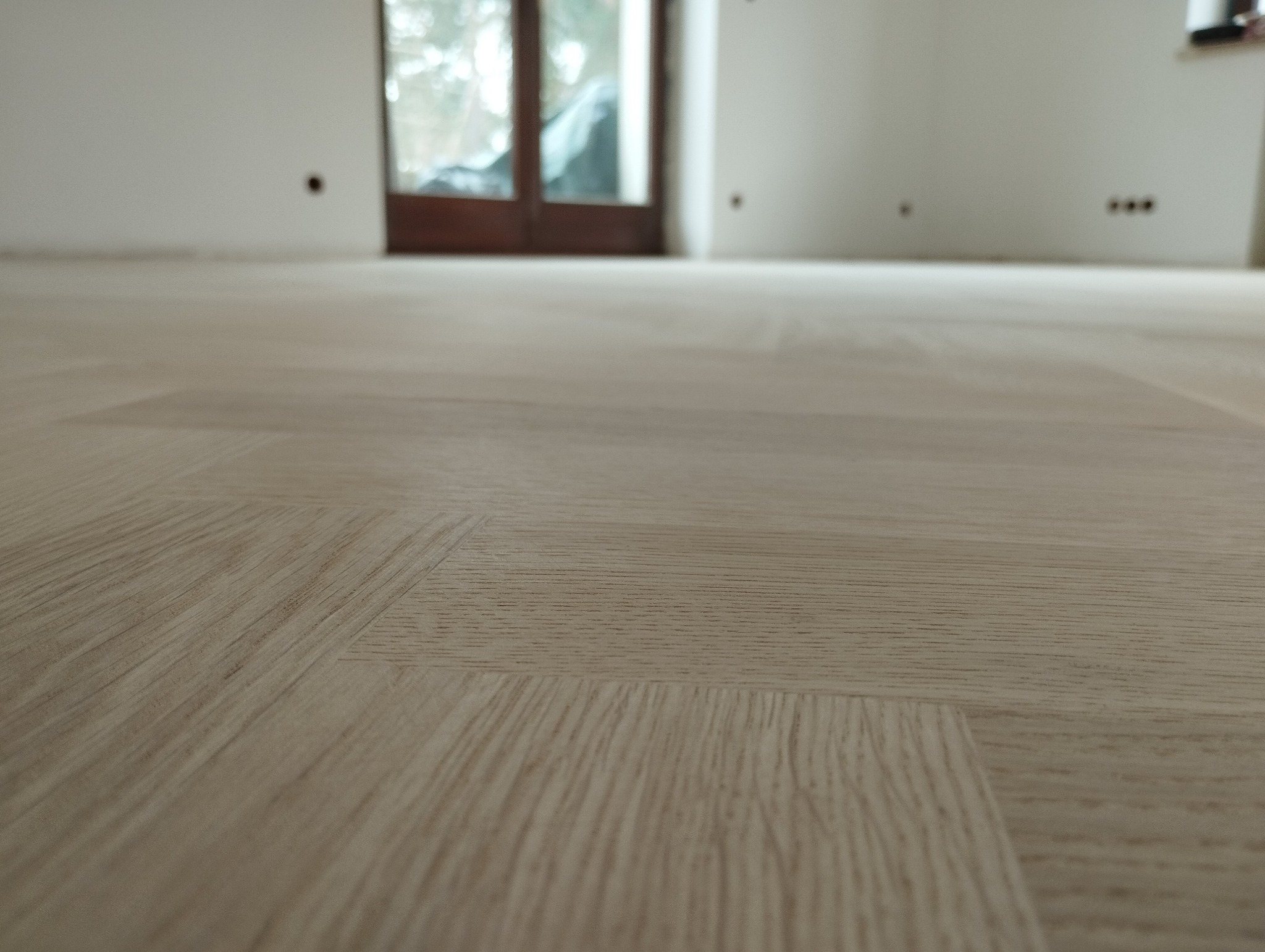 Jasna, nowo ułożona podłoga z paneli laminowanych z widocznym wzorem drewna, w tle drzwi balkonowe i ściany w stanie deweloperskim.