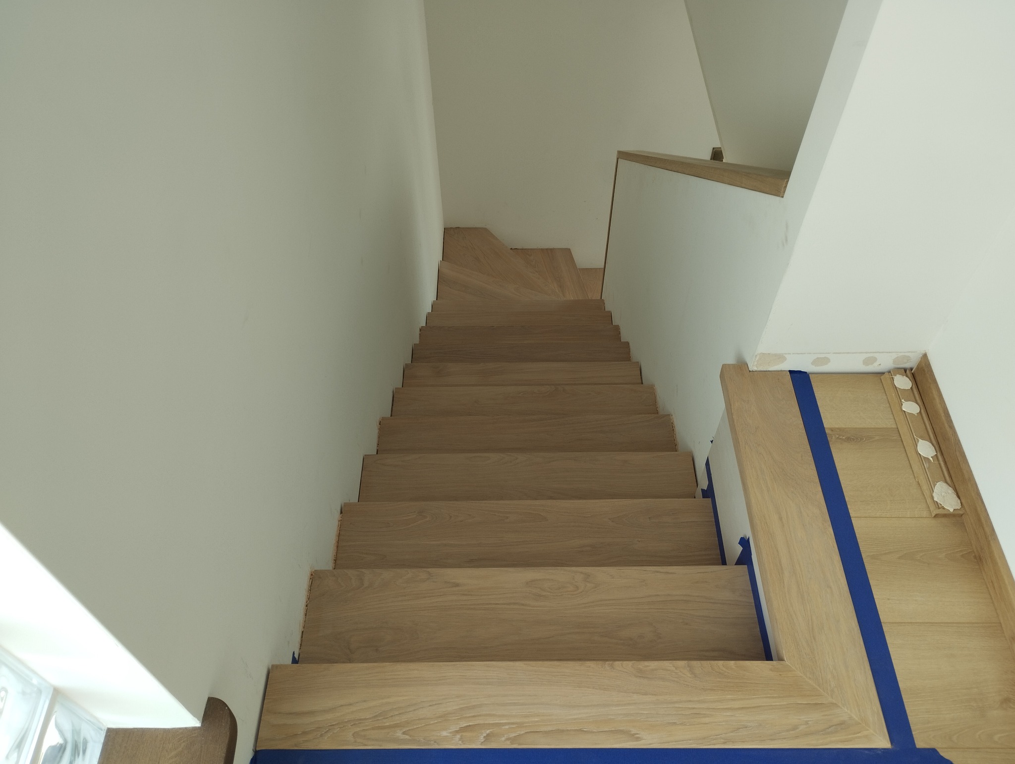 Widok z góry na jasne, drewniane schody z zabezpieczeniem w postaci niebieskiej taśmy malarskiej na podłodze i ścianie, widoczny fragment listwy przypodłogowej z wypełnieniami.