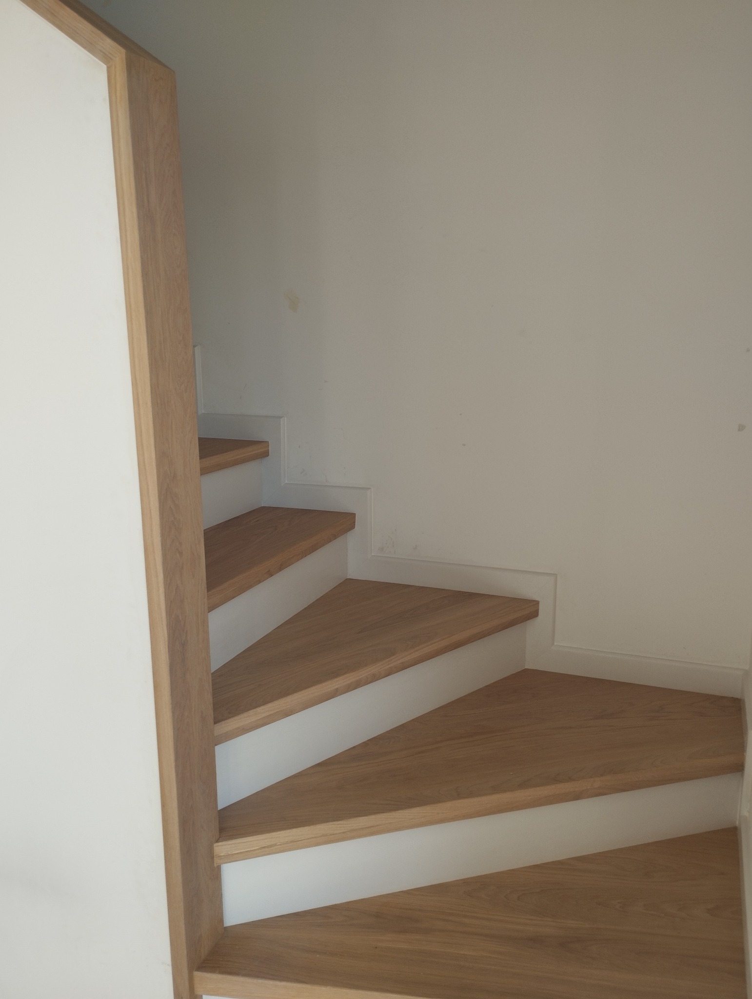 Wewnętrzne schody z drewnianymi stopniami i białymi podstopnicami, widoczne z dołu, z elementami ościeżnicy po lewej stronie.