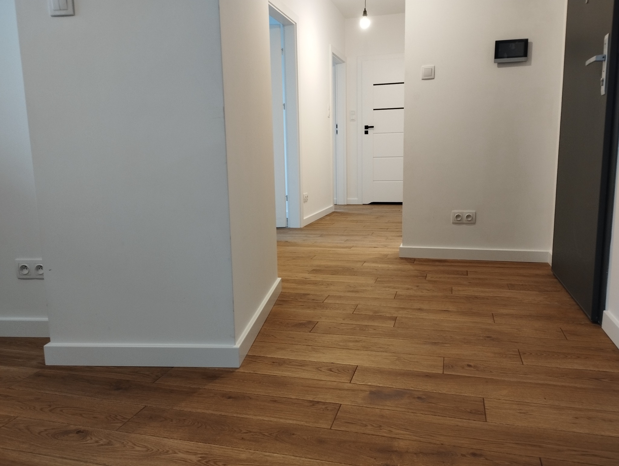 Ułożona podłoga z paneli drewnopodobnych w korytarzu z białymi ścianami, widoczne gniazdka elektryczne i białe listwy przypodłogowe.