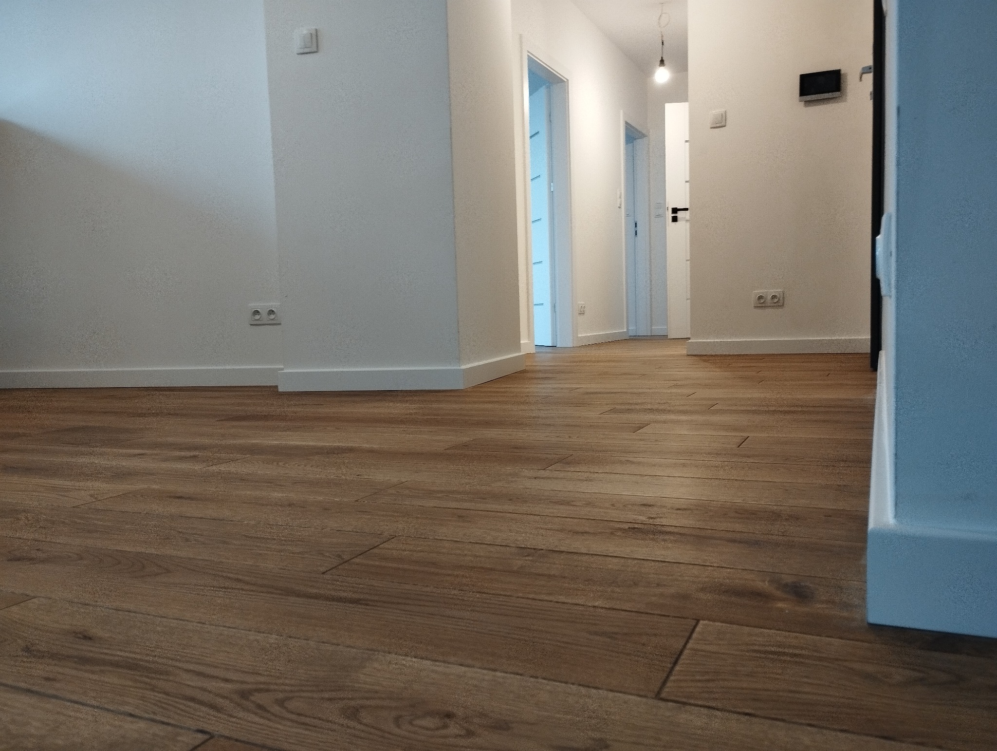 Ułożone panele podłogowe w odcieniach brązu, widok z niskiej perspektywy na korytarz z białymi ścianami i drzwiami.