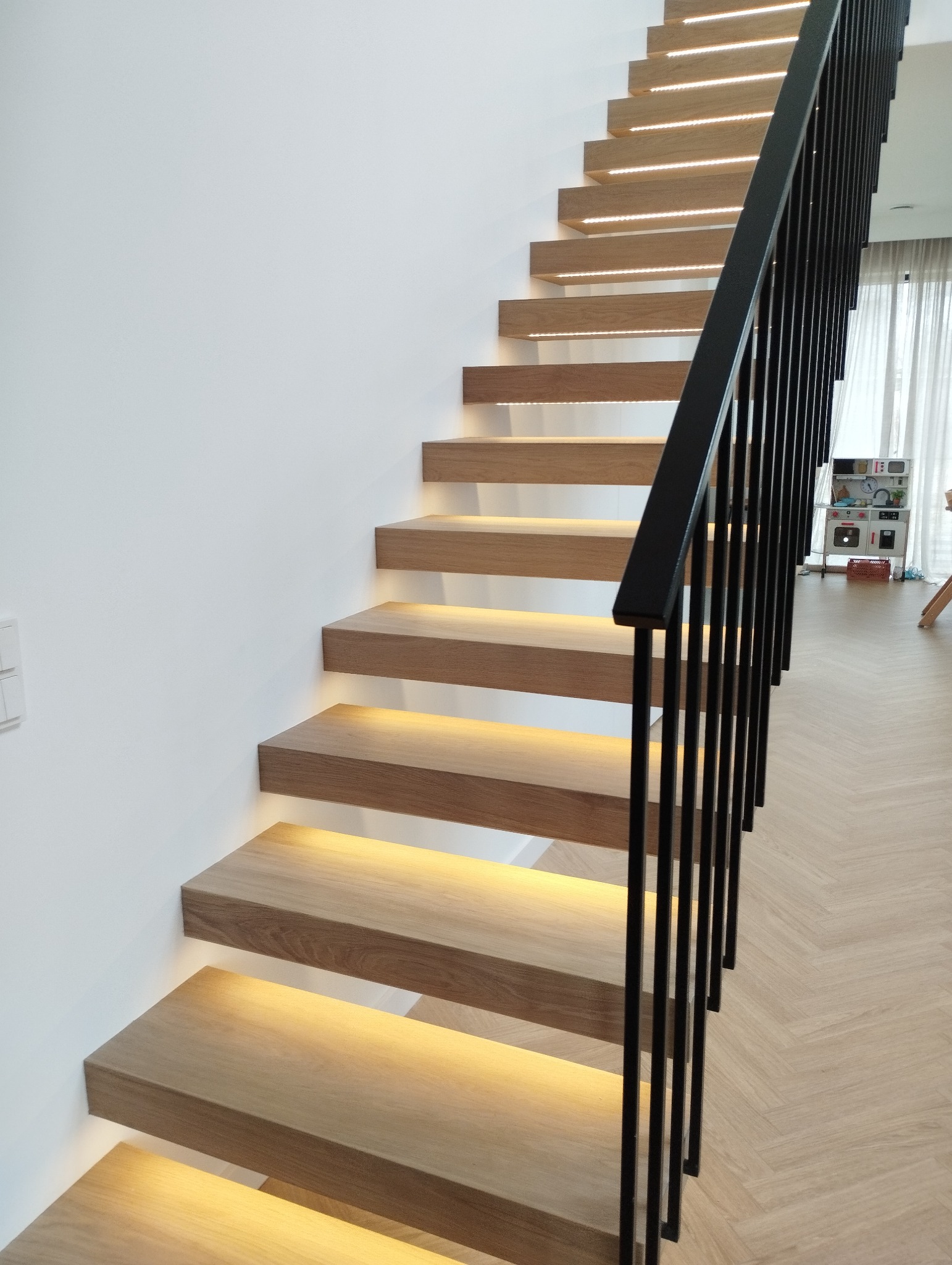 Nowoczesne schody z drewna z podświetleniem LED pod stopniami i czarną metalową balustradą, widok z dołu.