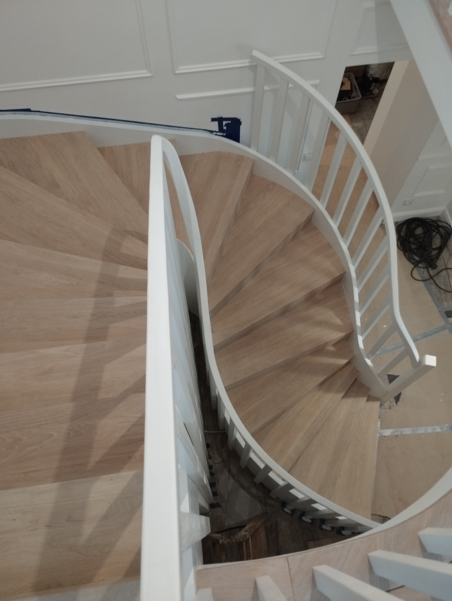Spiralne schody z jasnego drewna z białymi balustradami, widok z góry, w tle widoczne narzędzia i przewody.