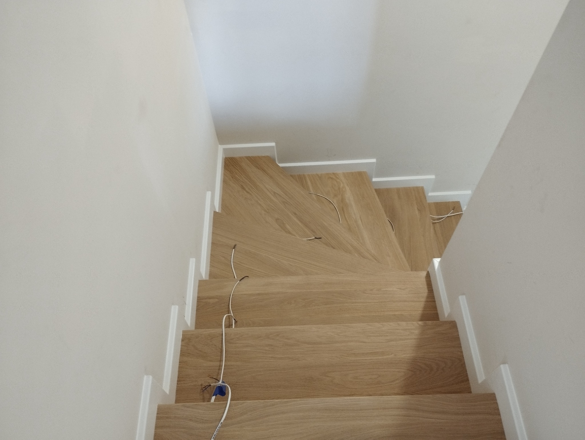 Jasne, dębowe schody z białymi listwami przypodłogowymi, widok z góry, z widocznymi przewodami elektrycznymi na kilku stopniach, sugerującymi trwający montaż oświetlenia.