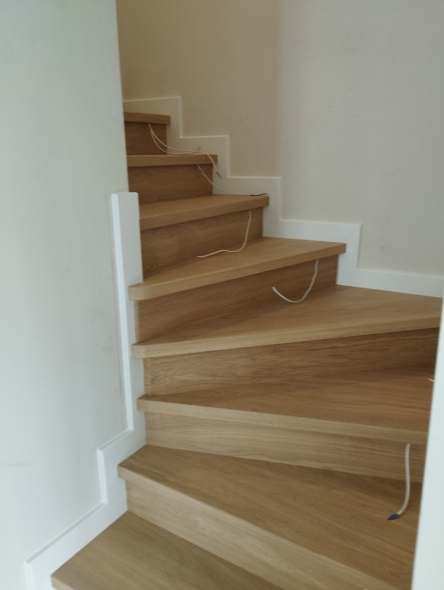 Wewnętrzne schody z jasnego drewna, z białymi listwami przypodłogowymi, widoczne niedociągnięcia w postaci wystających kabli.