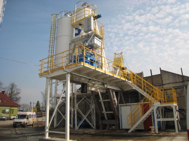 Kompletny węzeł betoniarski z dwoma silosami, schodami i żółtymi barierkami, widoczny z zewnątrz na tle nieba z chmurami i budynków.