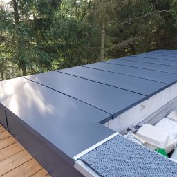 Producent - Rynny, Dachy, Obróbki blacharskie - Szary płaski dach z blachy trapezowej na budynku z drewnianym tarasem, widoczne drzewa w tle i białe bloczki budowlane.