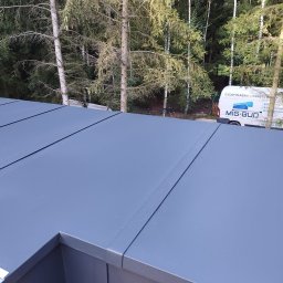 Producent - Rynny, Dachy, Obróbki blacharskie - Szare panele na dachu płaskim w trakcie montażu, widoczny fragment narzędzia i samochód firmy Mig-Bud w tle na tle drzew.