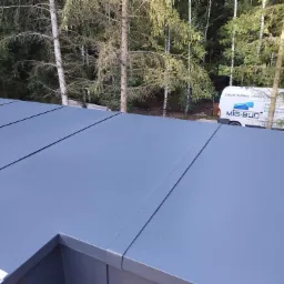 Szare panele na dachu płaskim w trakcie montażu, widoczny fragment narzędzia i samochód firmy Mig-Bud w tle na tle drzew.