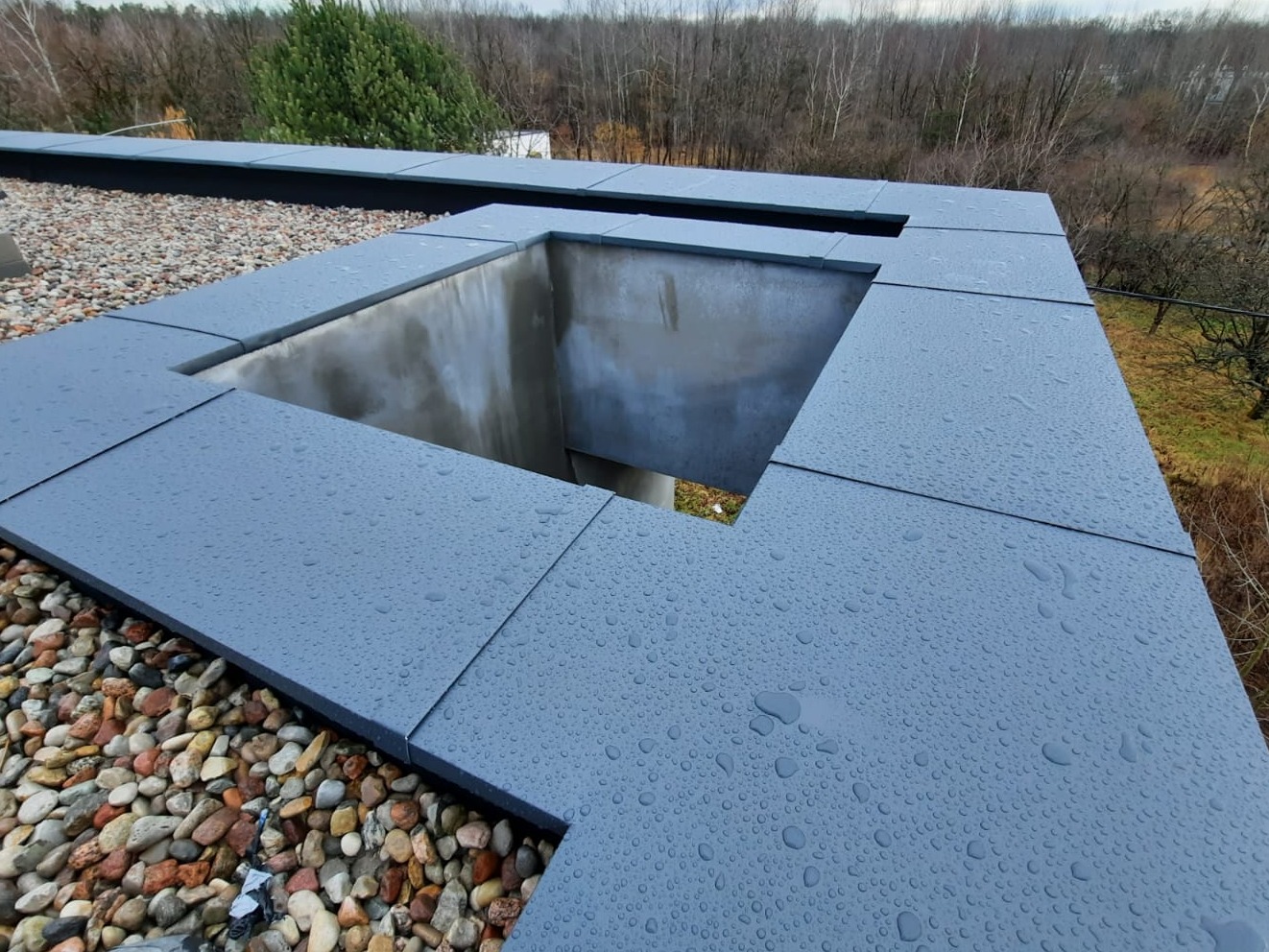 Płaski dach pokryty szarymi płytami z widocznym otworem wentylacyjnym i warstwą żwiru, pokryty kroplami deszczu.