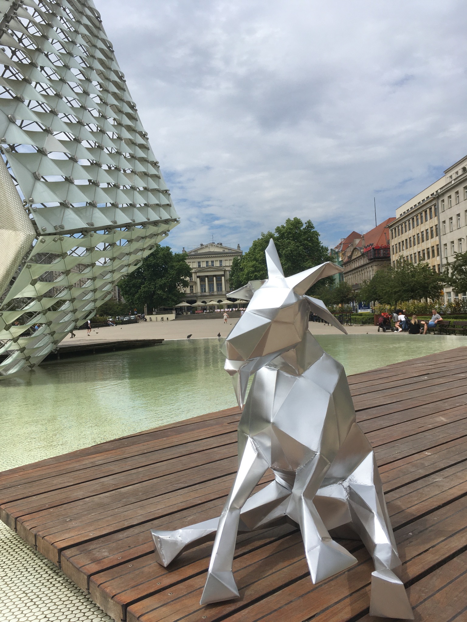 Geometryczna, metalowa rzeźba kozy siedzącej na drewnianym podeście obok nowoczesnej fontanny w kształcie sześcianu z trójkątnych paneli, w tle budynki miejskie i błękitne niebo z chmurami.