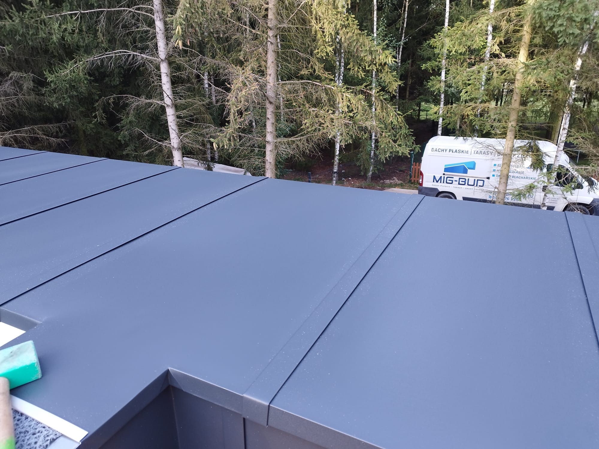 Szare panele na dachu płaskim w trakcie montażu, widoczny fragment narzędzia i samochód firmy Mig-Bud w tle na tle drzew.