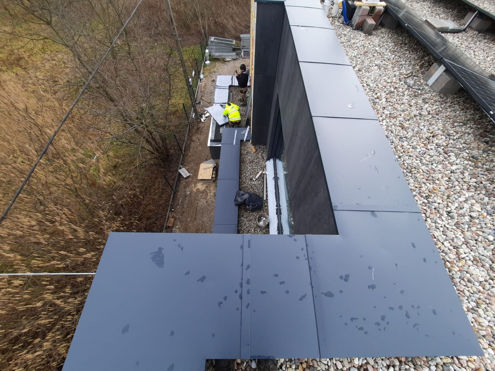 Montaż szarego opierzenia na dachu budynku, widoczne panele słoneczne, dwóch pracowników w trakcie pracy, jeden w żółtej kamizelce, otoczenie z kamieniami i roślinnością.