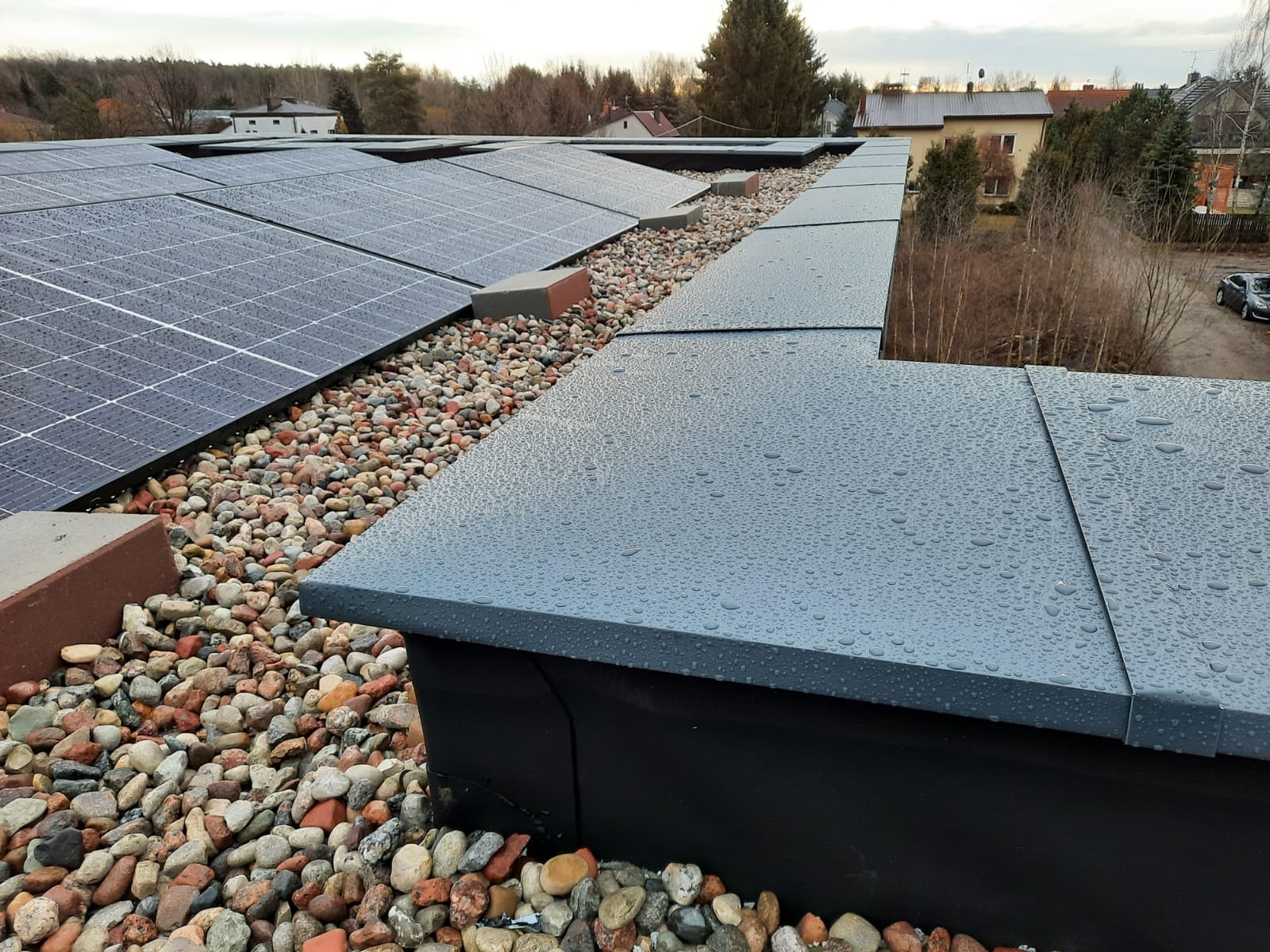 Płaski dach z panelami słonecznymi, pokryty drobnymi kamieniami i szarymi elementami wykończeniowymi, widoczne krople wody na powierzchni.
