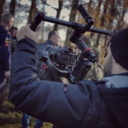 Operator filmowy trzymający profesjonalny stabilizator z kamerą Sony w plenerze, w tle grupa os&oacute;b i drzewa.