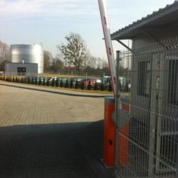 GERMAPLAN System Sp. z o.o. - Podniesiony szlaban automatyczny z pomarańczową obudową, biało-czerwonym ramieniem i metalową siatką ogrodzeniową w tle, widoczny budynek ochrony i zaparkowane samochody.