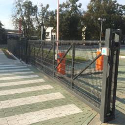 GERMAPLAN System Sp. z o.o. - Przesuwna brama z siatki metalowej, z automatycznym szlabanem i panelem sterowania na słupku, widoczna na tle chodnika z kostki brukowej i budynków.