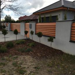 GERMAPLAN System Sp. z o.o. - Nowoczesne ogrodzenie posesji z poziomych, drewnopodobnych paneli w kolorze ciepłego brązu, osadzone na szarych, murowanych słupkach. Wzdłuż ogrodzenia posadzone małe, kuliste drzewka i krzewy...